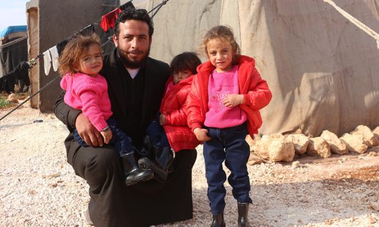 Man and three children wearing winter coats next to tents in Syria