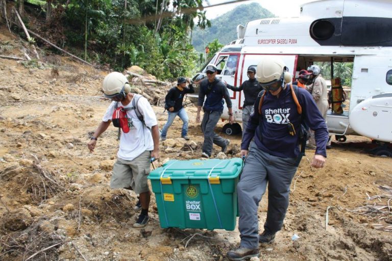 Two people wearing helmets carry a ShelterBox between them. in the background there is a helicopter.